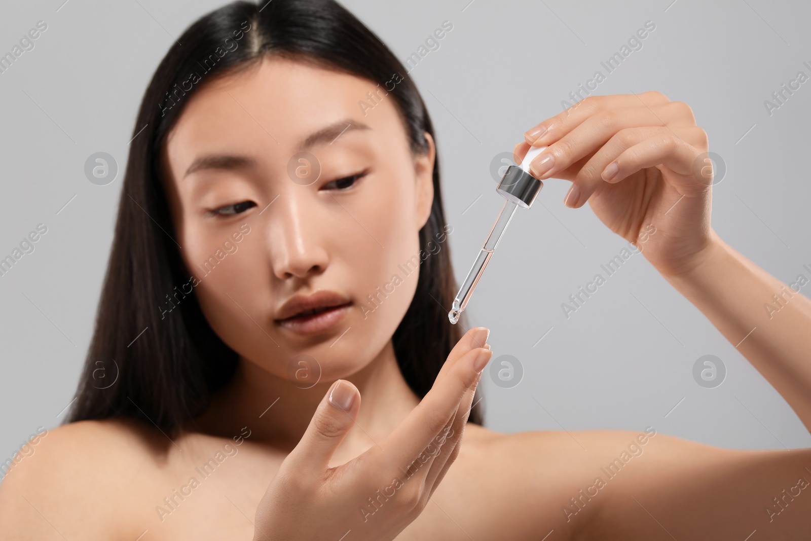 Photo of Beautiful young woman applying cosmetic serum onto her finger on grey background, selective focus