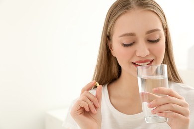Young woman with glass of water taking vitamin capsule on light background. Space for text