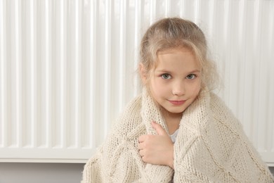 Photo of Little girl covered with knitted plaid sitting near heating radiator at home, space for text
