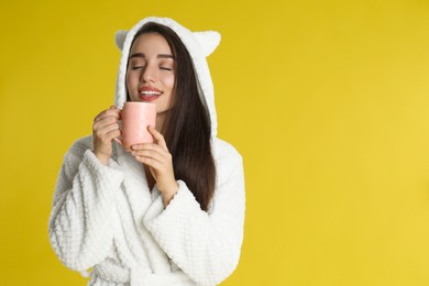 Young woman in bathrobe with cup of morning drink on yellow background. Space for text