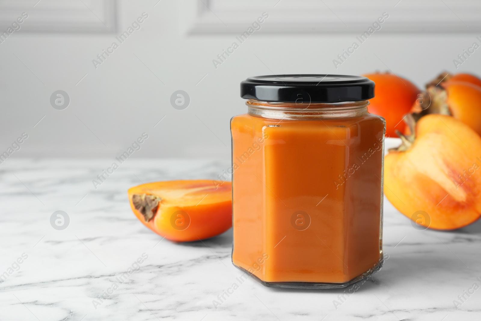 Photo of Delicious persimmon jam and fresh fruits on white marble table. Space for text