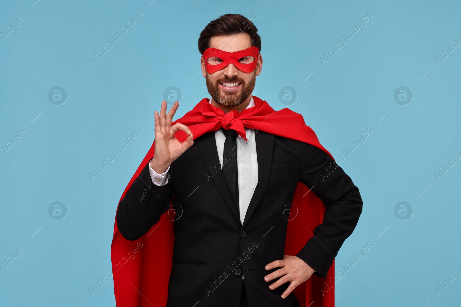 Photo of Businessman in red superhero cape and mask showing OK gesture on light blue background