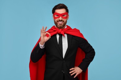 Photo of Businessman in red superhero cape and mask showing OK gesture on light blue background