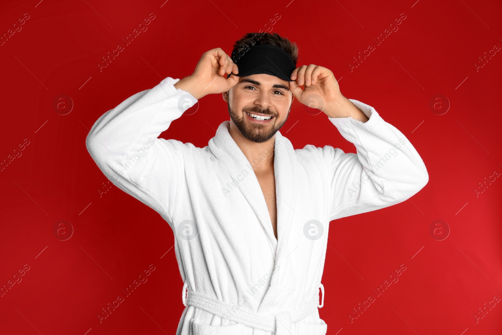 Photo of Happy young man in bathrobe and eye sleeping mask on red background