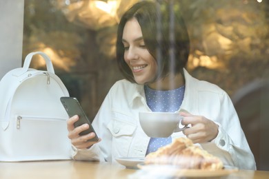Photo of Special Promotion. Happy young woman with cup of drink using smartphone in cafe, view from outdoors