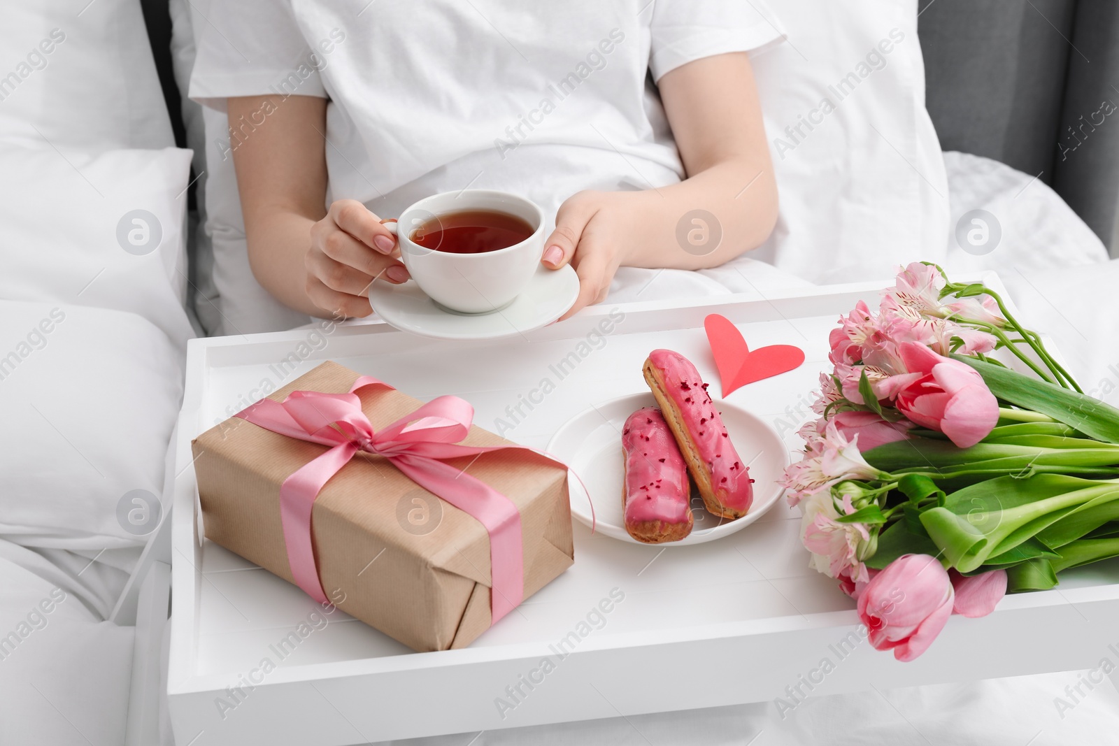 Photo of Tasty breakfast served in bed. Woman with tea, eclairs, gift box and flowers at home, closeup