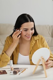 Photo of Beautiful young woman using eyelash curler while doing makeup at table indoors