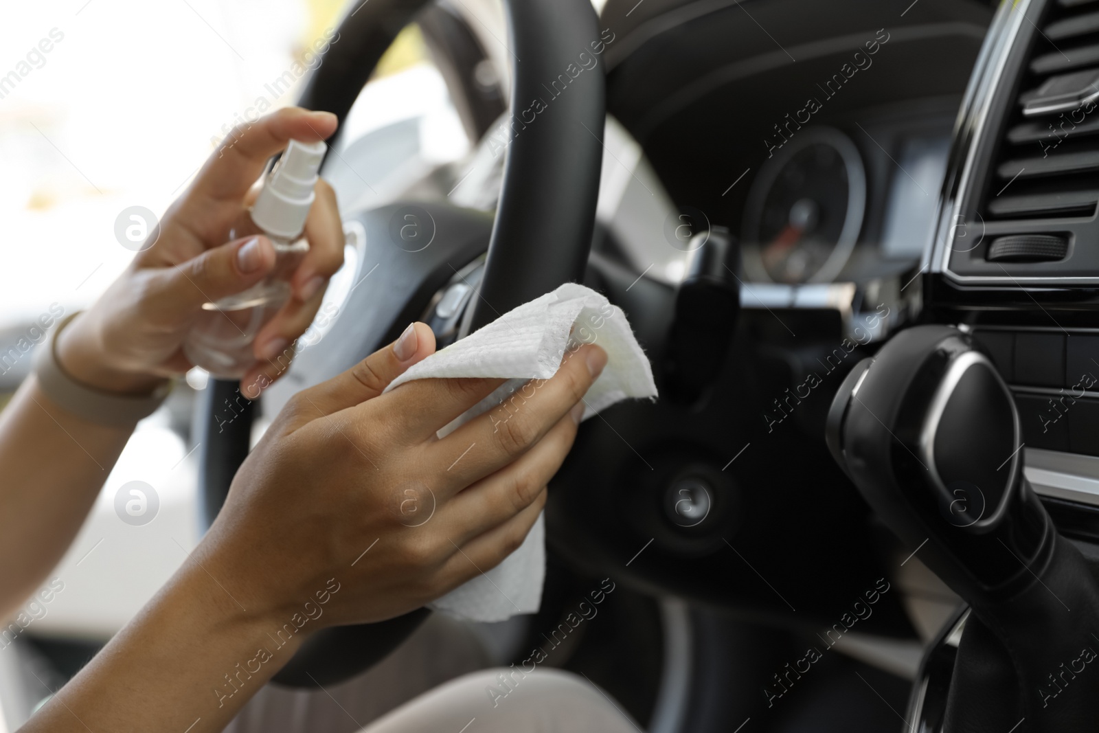 Photo of Woman cleaning steering wheel with wet wipe and antibacterial spray in car, closeup