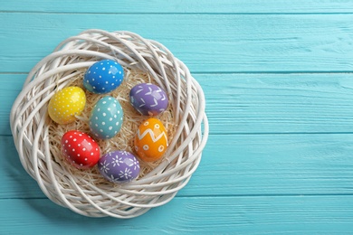 Wicker nest with painted Easter eggs on wooden table, top view. Space for text