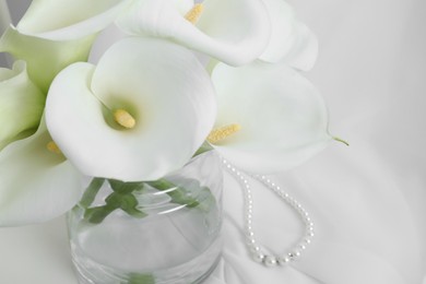 Photo of Beautiful calla lily flowers in glass vase on white cloth, closeup