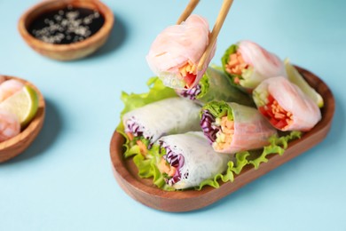Holding delicious spring roll with chopsticks on light blue background, closeup
