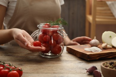 Woman putting tomatoes into glass jar at wooden kitchen table, closeup. Pickling vegetables