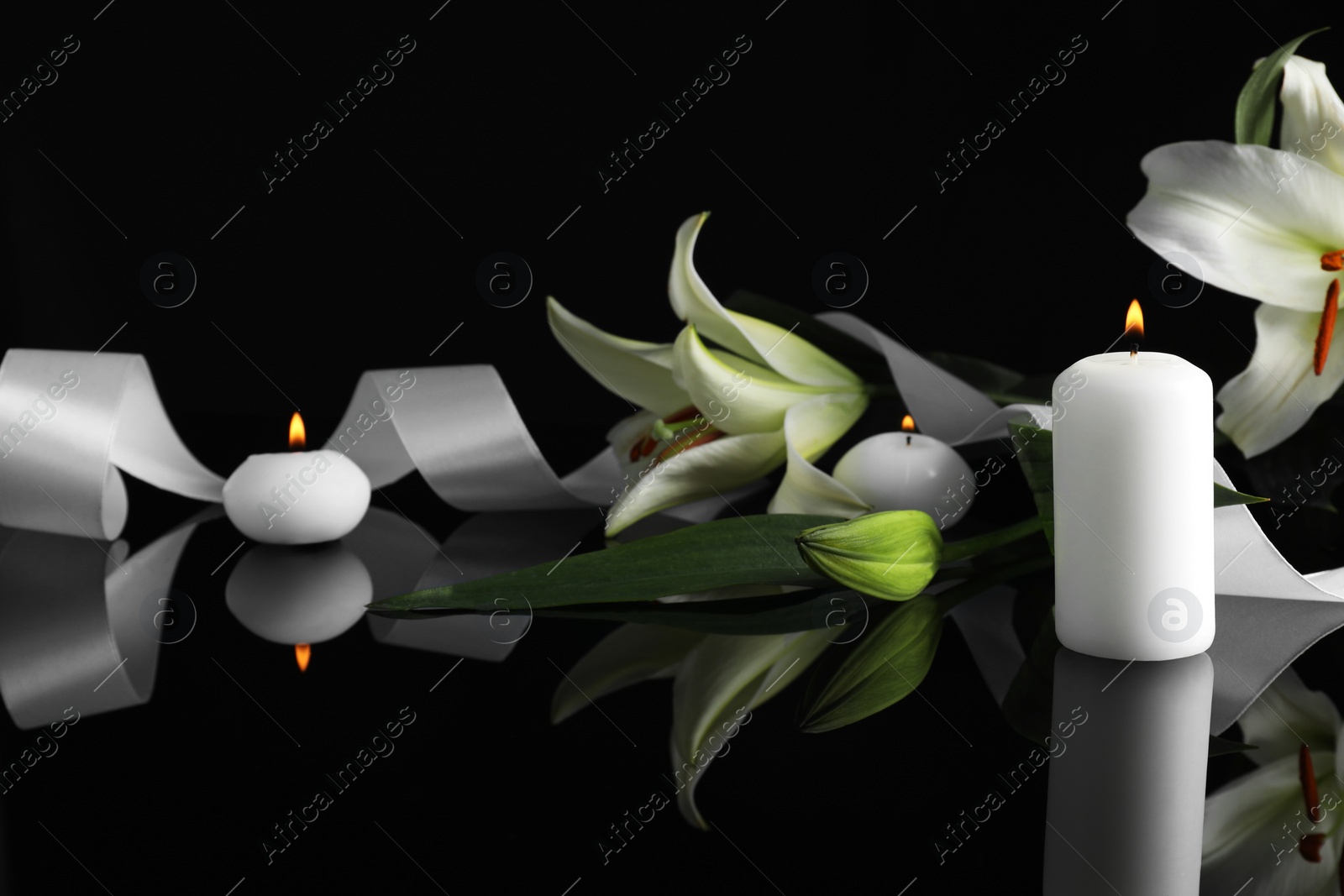 Photo of White lilies and burning candles on black mirror surface in darkness, space for text. Funeral symbols