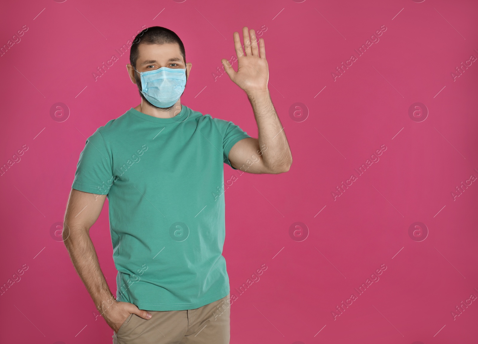 Photo of Man in protective mask showing hello gesture on pink background, space for text. Keeping social distance during coronavirus pandemic