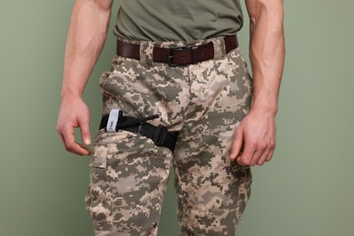 Man in military uniform with medical tourniquet on leg against light olive background, closeup