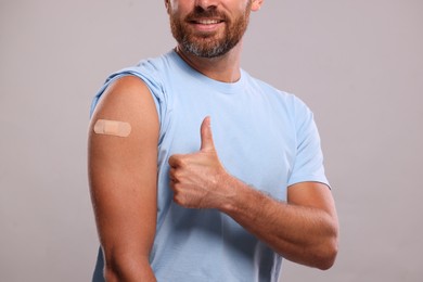 Photo of Man with sticking plaster on arm after vaccination showing thumbs up against light grey background, closeup