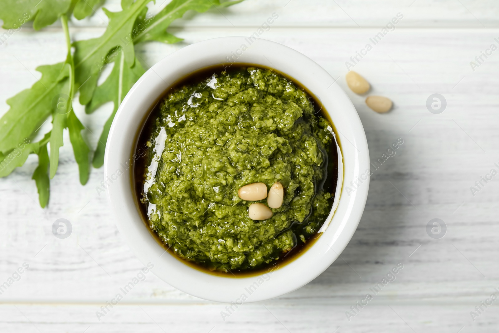 Photo of Bowl of tasty pesto and arugula on white wooden table, flat lay