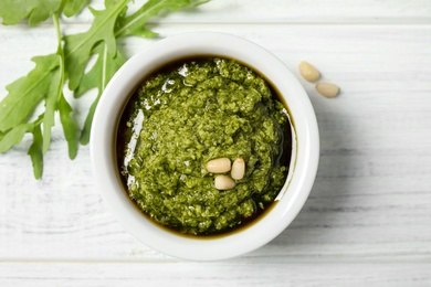 Bowl of tasty pesto and arugula on white wooden table, flat lay