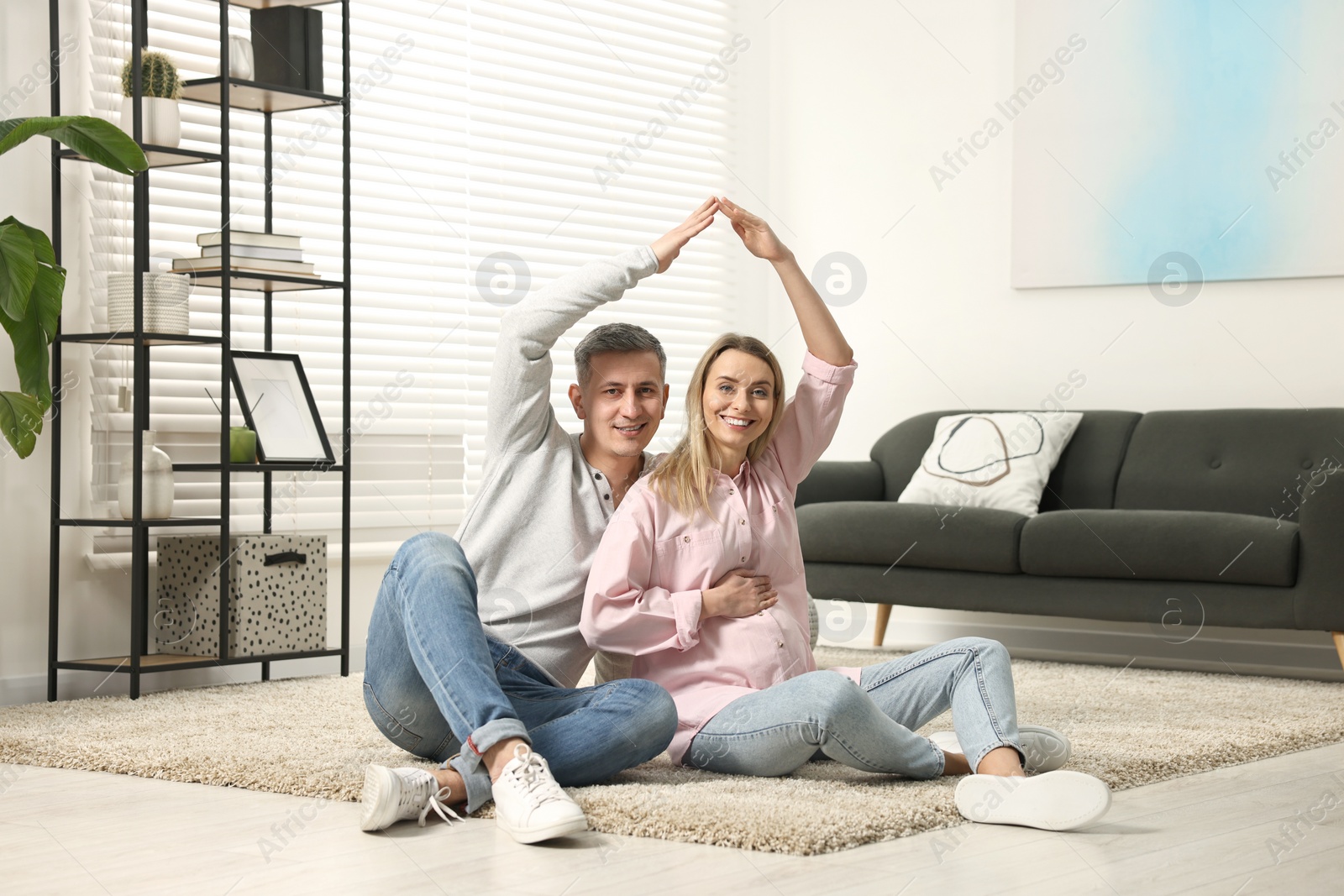 Photo of Young family housing concept. Pregnant woman with her husband forming roof with their hands while sitting on floor at home