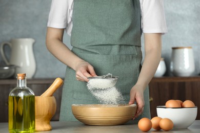 Photo of Woman sieving flour into bowl at table in kitchen, closeup