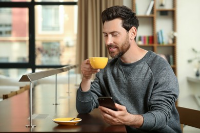 Handsome man with smartphone drinking coffee at table in cafe