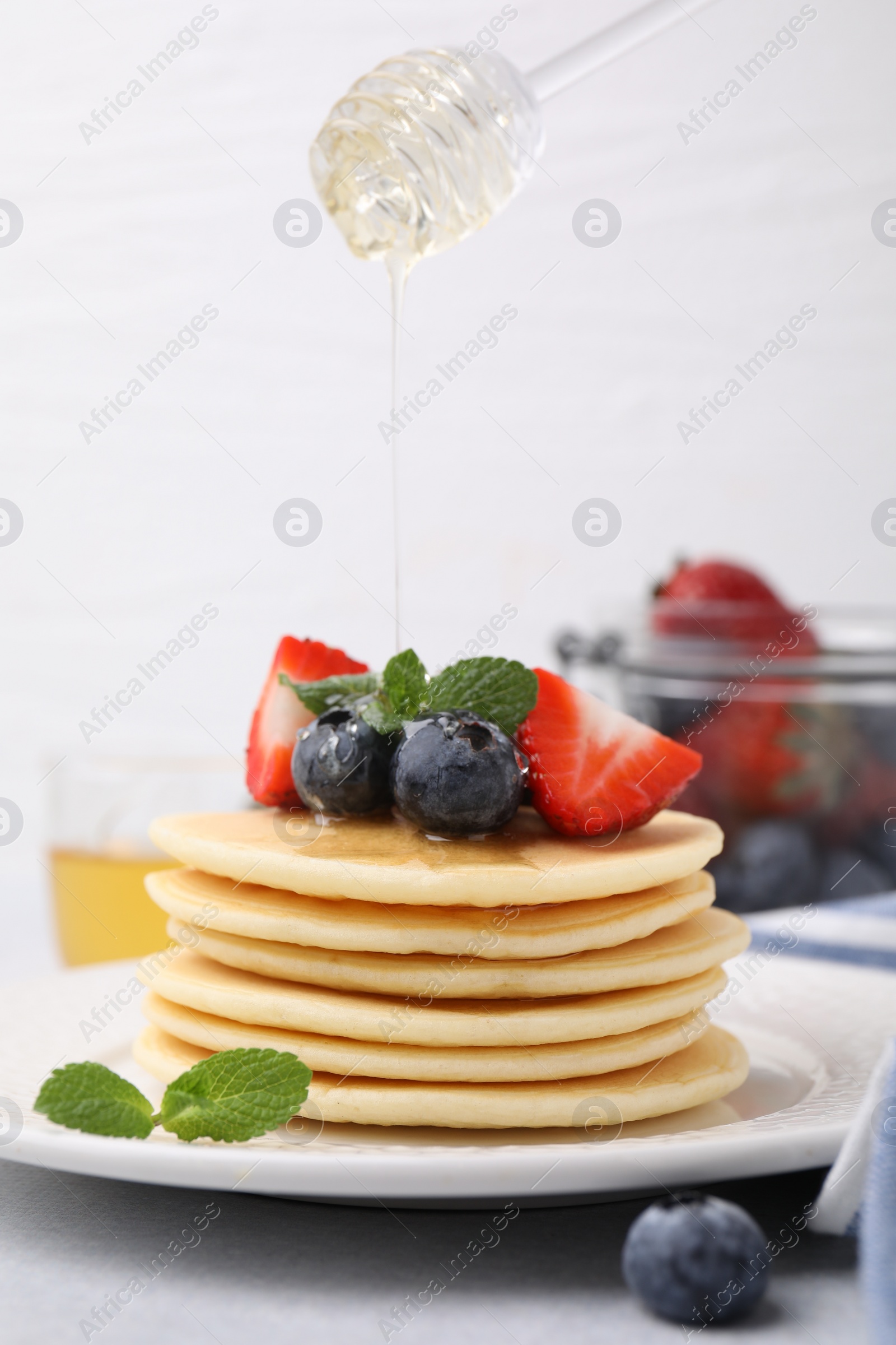 Photo of Pouring honey from dipper onto delicious pancakes with strawberries, blueberries and mint at light table, closeup