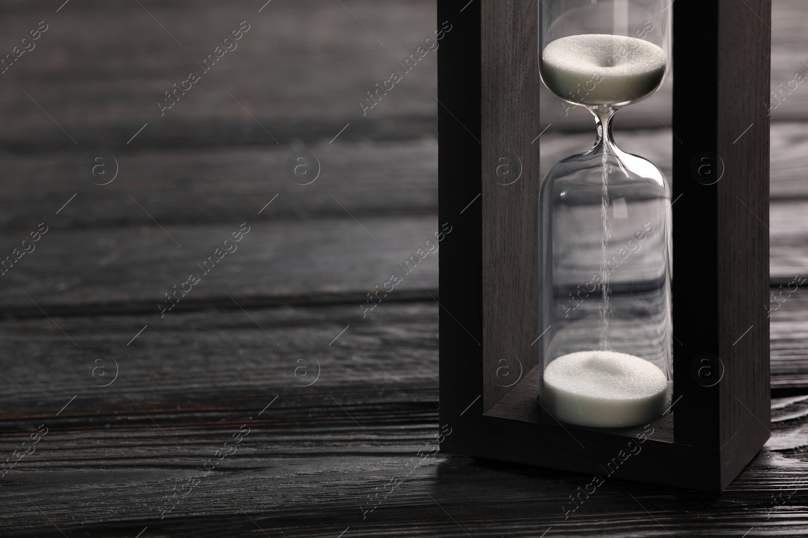 Photo of Hourglass with white flowing sand on black wooden table, closeup. Space for text