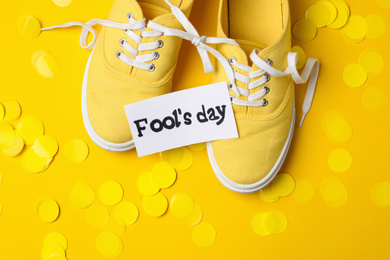 Photo of Shoes tied together, note with phrase FOOL'S DAY and confetti on yellow background, flat lay