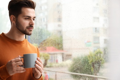 Photo of Thoughtful handsome man with cup of coffee near window indoors on rainy day