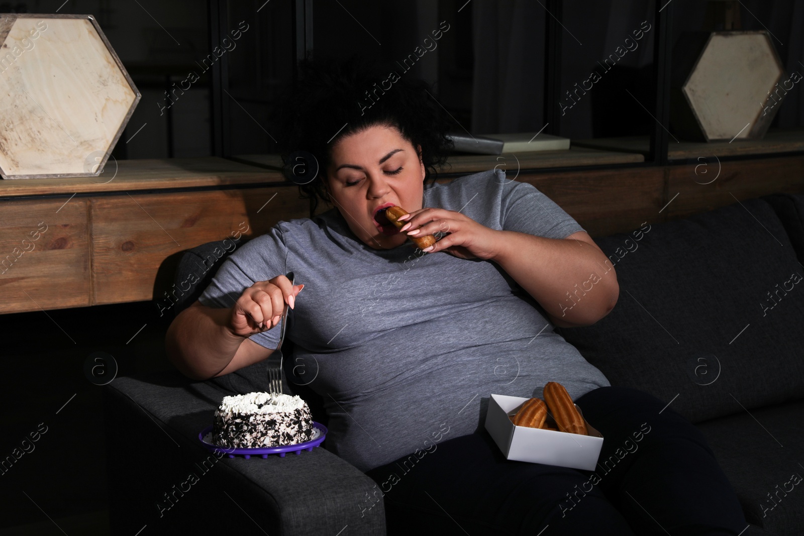 Photo of Depressed overweight woman eating sweets in living room at night