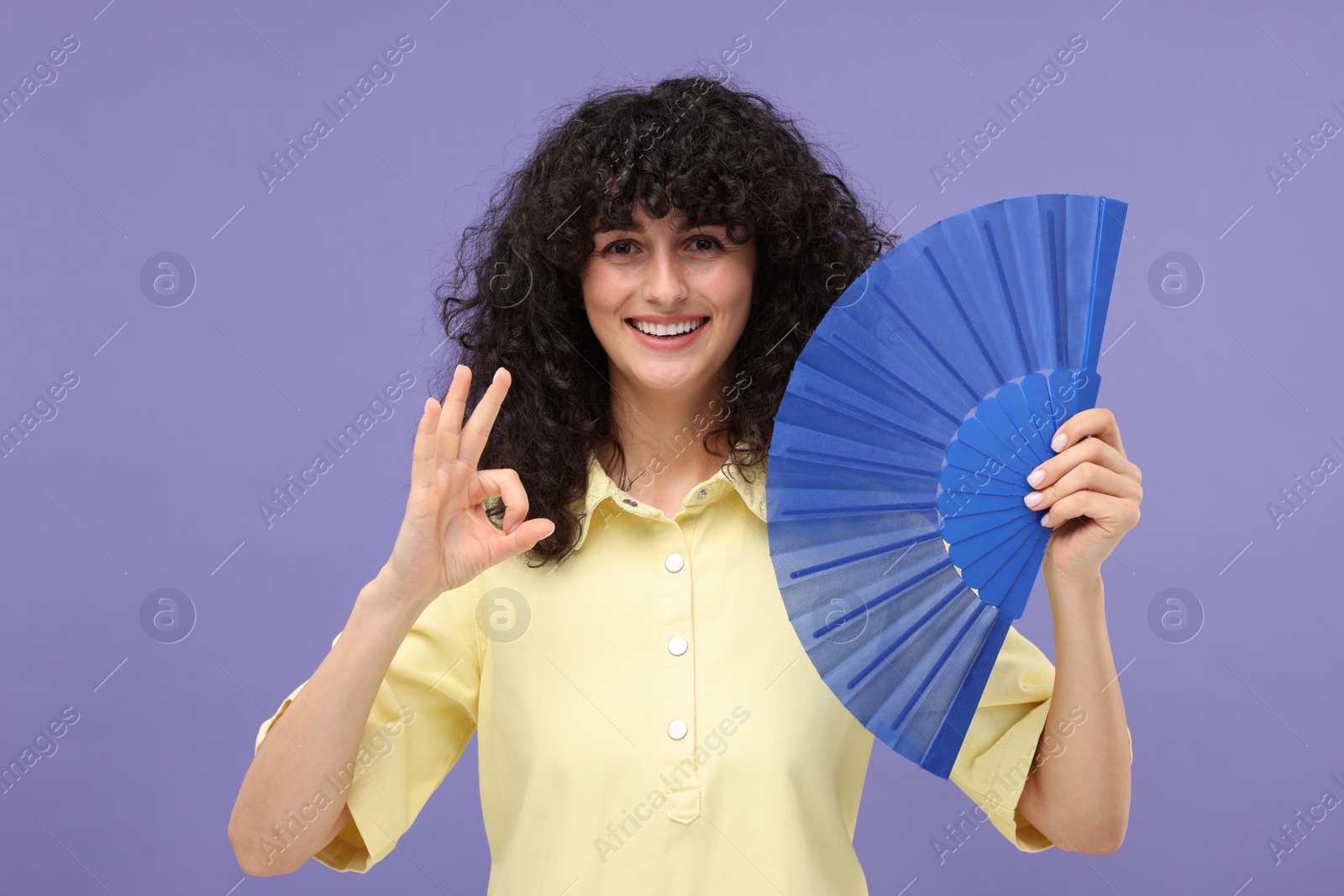 Photo of Happy woman holding hand fan and showing ok gesture on purple background