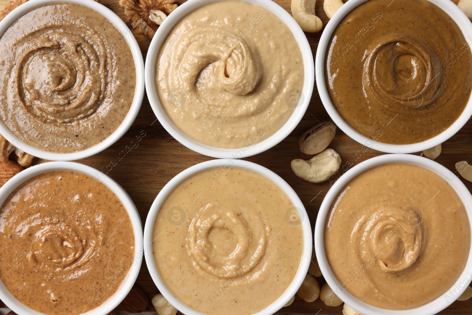 Photo of Many tasty nut butters in bowls and nuts on wooden table, flat lay