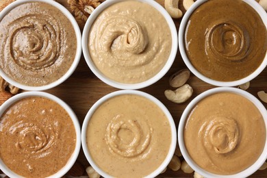 Many tasty nut butters in bowls and nuts on wooden table, flat lay