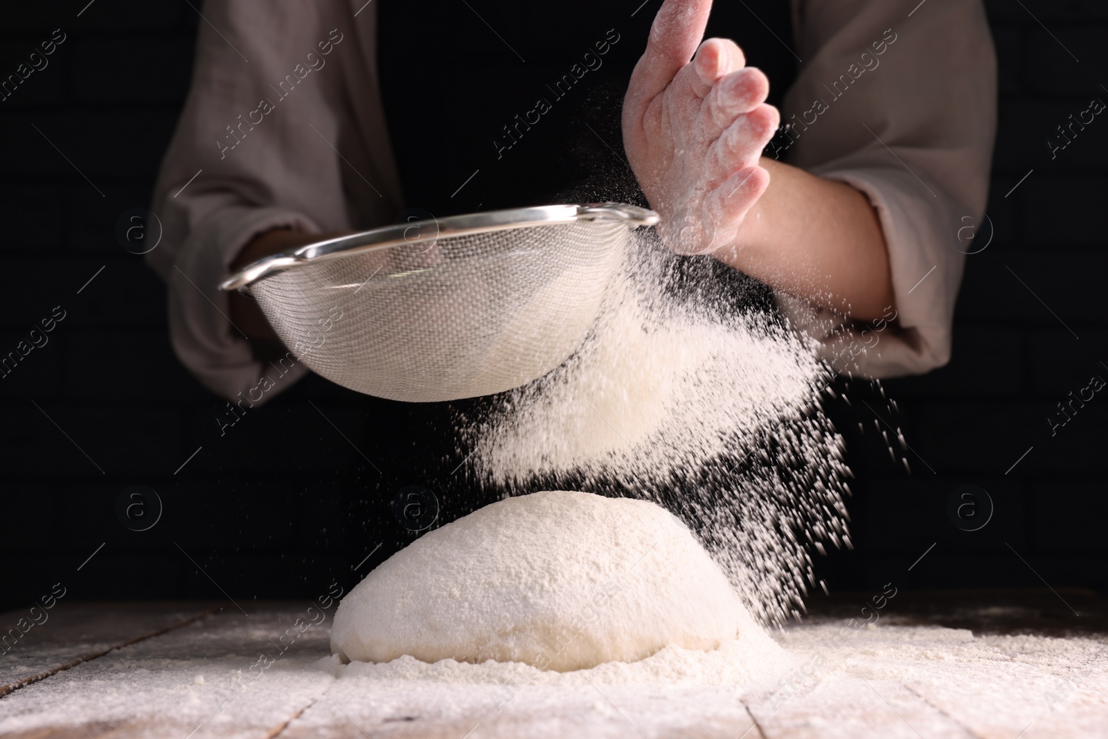 Photo of Woman sprinkling flour over dough at wooden table on dark background, closeup