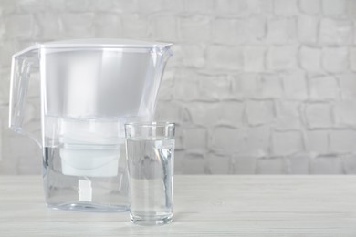 Photo of Filter jug and glass with purified water on white table indoors. Space for text