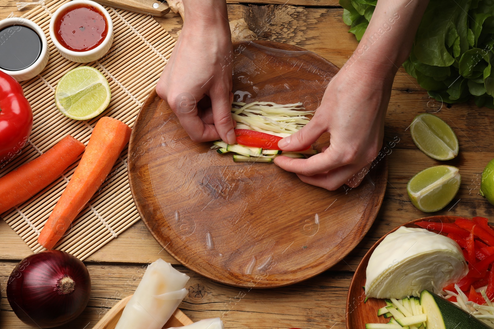 Photo of Making delicious spring rolls. Woman wrapping fresh vegetables into rice paper at wooden table, flat lay