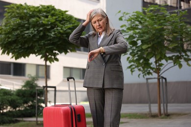 Photo of Being late. Worried senior businesswoman with red suitcase looking at watch outdoors