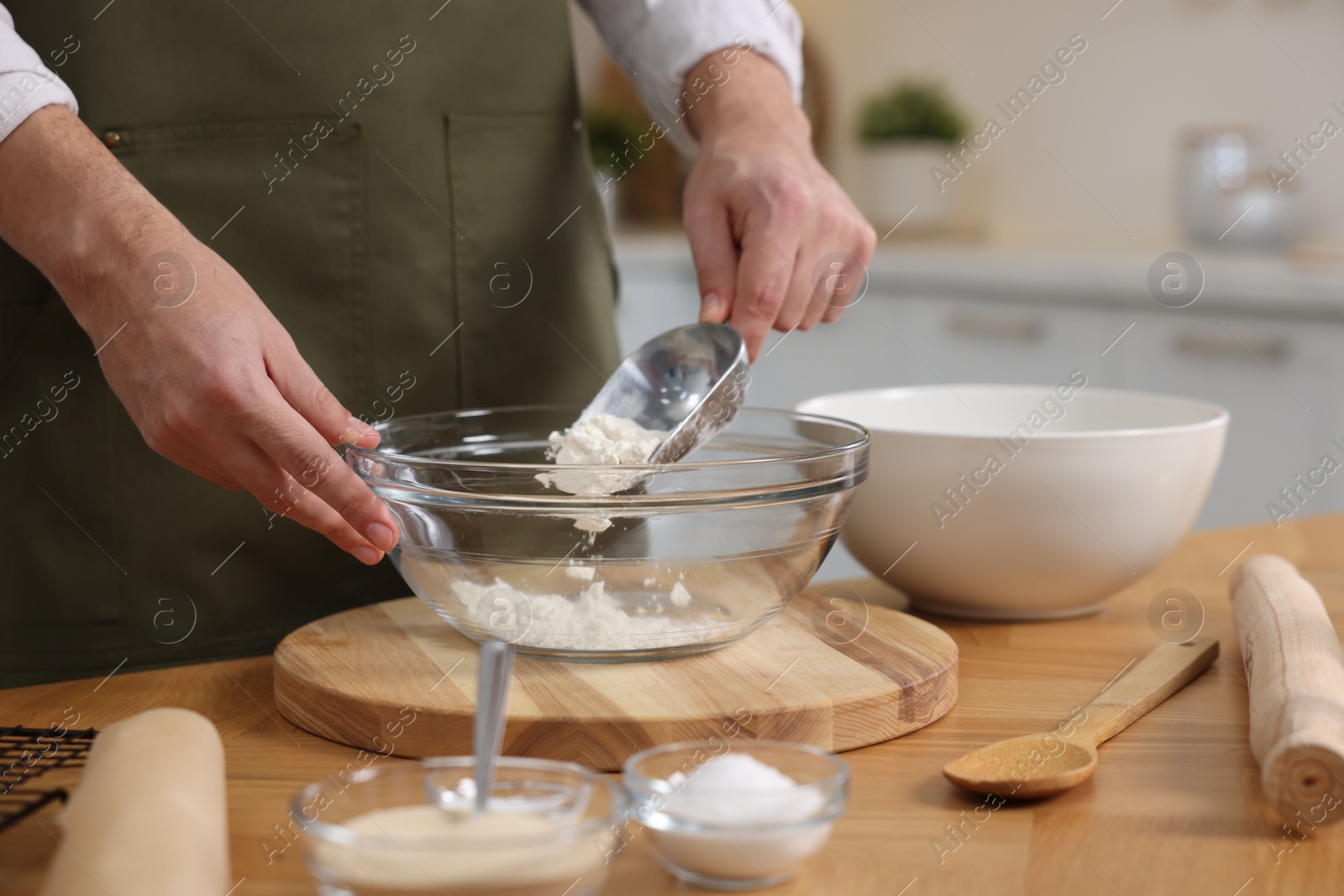 Photo of Making bread. Man putting flour into bowl at wooden table in kitchen, closeup