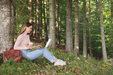Young woman with cup of coffee working on laptop in forest, space for text