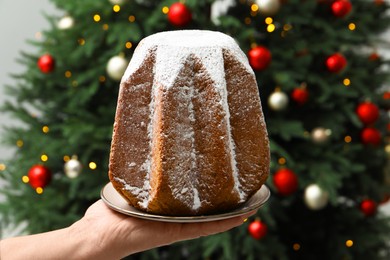 Photo of Woman holding delicious Pandoro cake decorated with powdered sugar near Christmas tree, closeup. Traditional Italian pastry