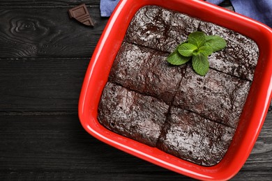 Delicious chocolate brownie with mint in baking dish on black wooden table, flat lay. Space for text