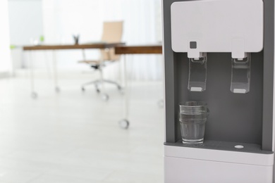 Photo of Modern water cooler with glass in office, closeup. Space for text