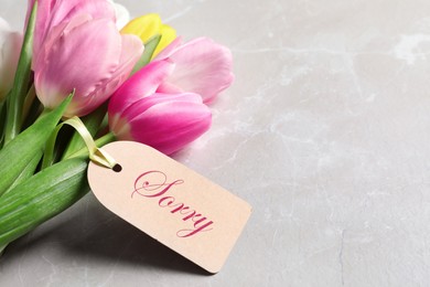 Image of Word Sorry written on label and bouquet of tulips on light grey marble table, closeup. Space for text