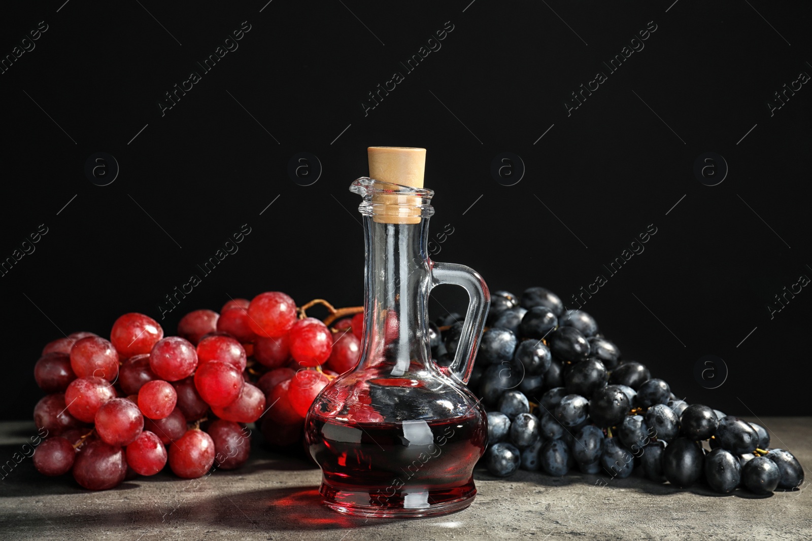 Photo of Glass jug with wine vinegar and fresh grapes on gray table against black background