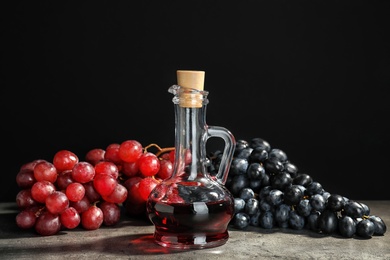 Photo of Glass jug with wine vinegar and fresh grapes on gray table against black background