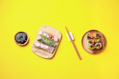 Photo of Different delicious spring rolls, chopsticks and soy sauce on yellow background, flat lay