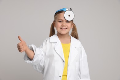 Photo of Little girl in medical uniform with head mirror showing thumb up on light grey background