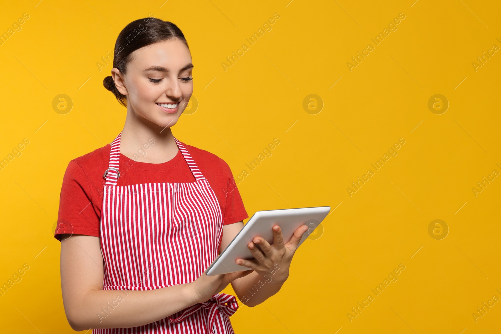 Photo of Beautiful young woman in clean striped apron with tablet on orange background. Space for text