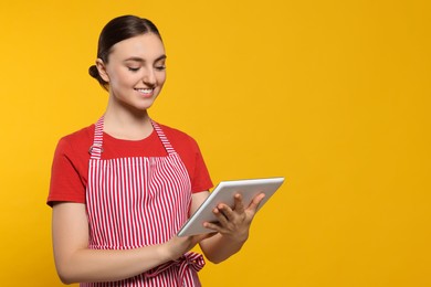 Photo of Beautiful young woman in clean striped apron with tablet on orange background. Space for text