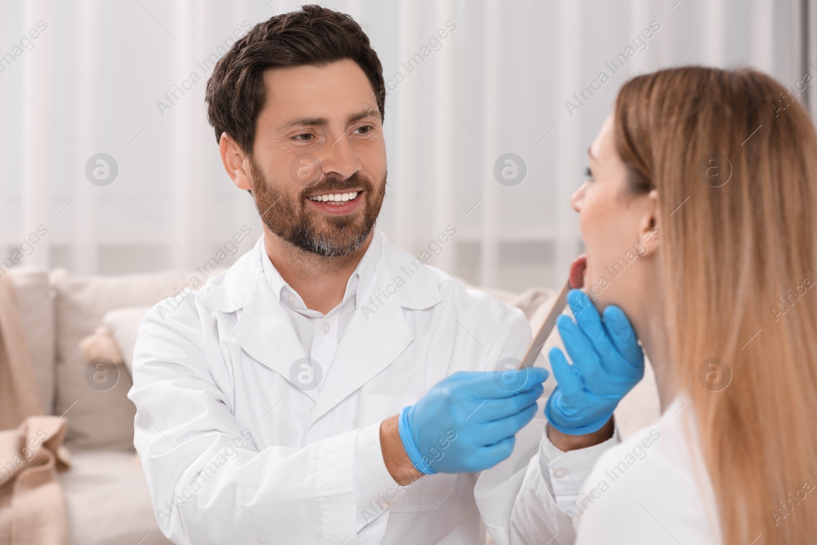 Photo of Smiling doctor examining woman`s oral cavity with tongue depressor indoors